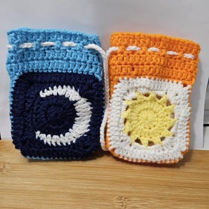 May include: Two small crochet drawstring pouches. The left pouch is light blue with a dark blue square featuring a white crescent moon. The right pouch is orange with a white square featuring a yellow sun. Both have white drawstring cords.