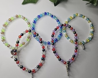 Puede incluir: Cinco pulseras circulares de cuentas dispuestas en un patrón superpuesto. Las pulseras presentan cuentas de vidrio translúcidas y multicolores en tonos azul, verde, rojo, amarillo y morado, con dijes de metal plateado, incluyendo una espada.