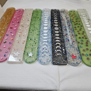 May include: A collection of nine rectangular resin incense holders arranged in a row. Each holder features unique embedded elements including glitter, stars, moon phases, flowers, and small colorful shapes in various colors like gold, pink, green, blue, and black.
