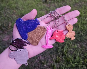 Puede incluir: Una colección de siete llaveros con diversas figuras esculpidas, incluyendo un busto azul, un rostro dorado, una sirena rosa, una forma abstracta roja, un busto pequeño dorado, una forma morada oscura y un busto de perfil gris claro en un cordón.