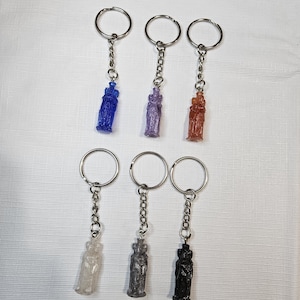 May include: Six keychains featuring small, upright, molded figures attached to silver-toned metal split rings by short chains. The figures are translucent and opaque in blue, purple, copper, clear, silver, and black, each with a distinct textured surface.