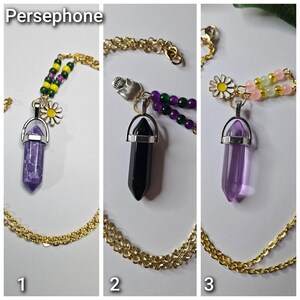 Puede incluir: Tres collares de cadena dorada con colgantes de cristal en punta. El collar 1 tiene una piedra morada, cuentas verdes y amarillas y un dije de margarita. El collar 2 tiene una piedra negra, cuentas moradas y verdes y un dije de calavera. El collar 3 tiene una piedra morada transparente, cuentas rosas y amarillas y un dije de margarita.