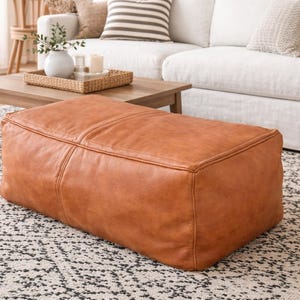 May include: A rectangular, brown leather ottoman sits on a patterned rug. The ottoman is a warm, caramel color with visible stitching details. The room features a white sofa, wooden coffee table, and decorative items.