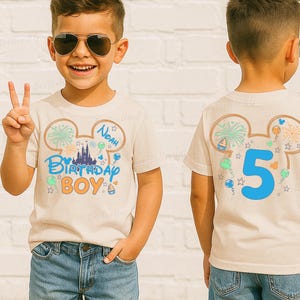 Op de afbeelding: Een beige t-shirt met een kleurrijk verjaardagsontwerp. De voorkant heeft de tekst "Birthday Boy" en de naam "Noah". De achterkant toont het nummer "5". Het ontwerp bevat vuurwerk, ballonnen en een kasteel.