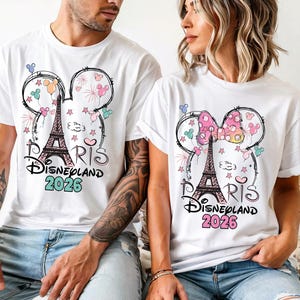 May include: White t-shirts with a graphic design featuring the Eiffel Tower, Mickey Mouse ears, and the text "Paris Disneyland 2026". The design includes colorful accents like pink, blue, and green.