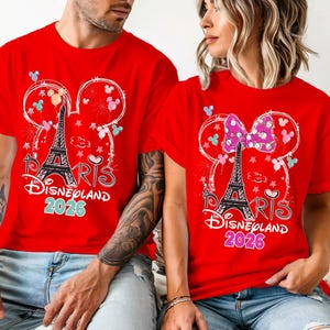 May include: Red t-shirts with a graphic featuring the Eiffel Tower, Mickey Mouse ears, and the text "Paris Disneyland 2026." One shirt has a bow. The shirts are worn by two people.