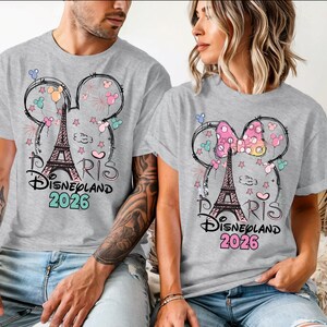 May include: Gray t-shirts featuring a design with the Eiffel Tower, Mickey Mouse ears, and the text "Paris Disneyland 2026." The design includes colorful balloons, stars, and fireworks. The shirts are worn by two people.