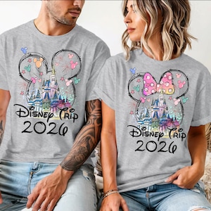K&ouml;nnte beinhalten: Zwei graue T-Shirts mit einem farbenfrohen Disney-Schloss-Design in einer Mickey-Mouse-Kopf-Umrisslinie. Die Shirts tragen in schwarzer Schrift "Disney Trip 2026". Ein Shirt hat eine rosa Schleife, das andere ein Mickey-Mouse-Ohren-Design.