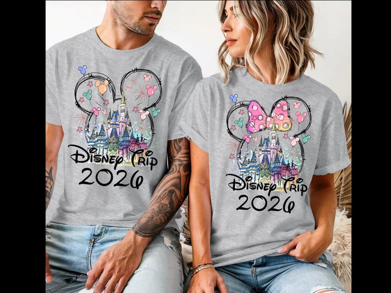 K&ouml;nnte beinhalten: Zwei graue T-Shirts mit einem farbenfrohen Disney-Schloss-Design in einer Mickey-Mouse-Kopf-Umrisslinie. Die Shirts tragen in schwarzer Schrift "Disney Trip 2026". Ein Shirt hat eine rosa Schleife, das andere ein Mickey-Mouse-Ohren-Design.