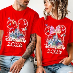 K&ouml;nnte beinhalten: Rote T-Shirts mit einem Disney Trip 2026 Design. Das Design zeigt ein Schloss, Feuerwerk und Mickey Mouse Ohren. Ein Shirt hat eine Schleife auf den Ohren. Die Shirts werden von zwei Personen getragen.