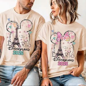 May include: Beige t-shirts featuring a Mickey Mouse head design with the Eiffel Tower, fireworks, and stars. The text "Paris Disneyland 2026" is printed below the design. One shirt has a bow.