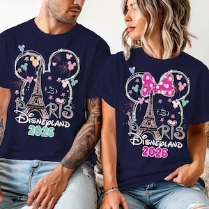 May include: Navy blue t-shirts with a Disneyland Paris 2026 design. The design includes the Eiffel Tower, Mickey Mouse ears, fireworks, and the word "Paris" in a script font. One shirt has a pink bow.