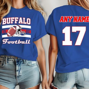 Puede incluir: Dos camisetas azul real con detalles en blanco y rojo. La camiseta delantera tiene el texto "BUFFALO" con un casco de fútbol y el texto "Football". La camiseta trasera tiene el texto "ANY NAME" y el número "17".
