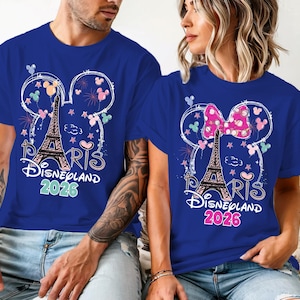May include: Two blue t-shirts with a Disneyland Paris 2026 design. The design includes the Eiffel Tower, Mickey Mouse ears, fireworks, and the word "Paris". One shirt has a pink bow.