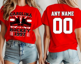Puede incluir: Dos camisetas rojas con texto y gráficos blancos. Una camiseta dice "Carolina Hockey 1997" con un palo de hockey y dos patines de hielo. La otra camiseta dice "Any Name 00".