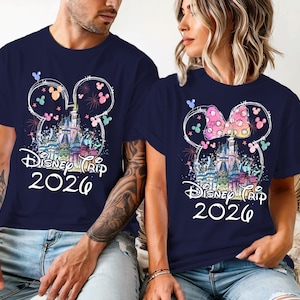 K&ouml;nnte beinhalten: Marineblaue T-Shirts mit einem farbenfrohen Disney-Schloss-Design, Mickey Mouse Ohren und dem Text "Disney Trip 2026". Ein Shirt hat eine rosa Schleife. Die Shirts werden von zwei Personen getragen.