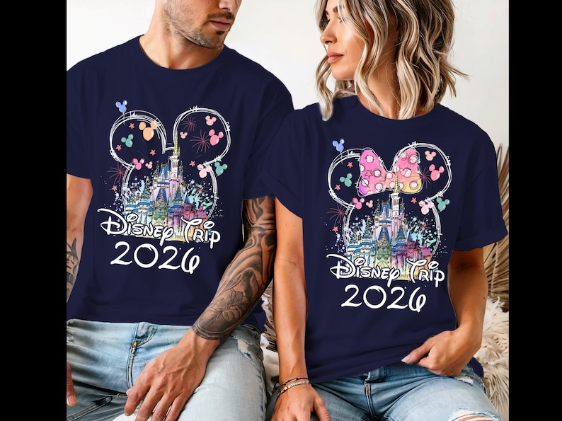 K&ouml;nnte beinhalten: Marineblaue T-Shirts mit einem farbenfrohen Disney-Schloss-Design, Mickey Mouse Ohren und dem Text "Disney Trip 2026". Ein Shirt hat eine rosa Schleife. Die Shirts werden von zwei Personen getragen.