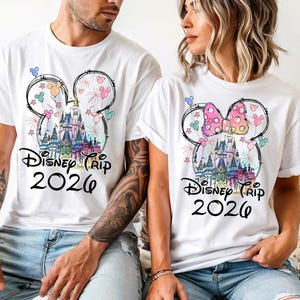 K&ouml;nnte beinhalten: Wei&szlig;e T-Shirts mit einem farbenfrohen Disney-Schloss-Motiv in einer Mickey-Mouse-Kopf-Umrisslinie. Der Text "Disney Trip 2026" ist darunter gedruckt. Ein Shirt hat eine rosa Schleife.