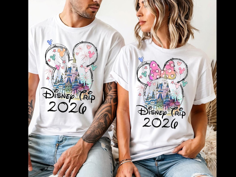 K&ouml;nnte beinhalten: Wei&szlig;e T-Shirts mit einem farbenfrohen Disney-Schloss-Motiv in einer Mickey-Mouse-Kopf-Umrisslinie. Der Text "Disney Trip 2026" ist darunter gedruckt. Ein Shirt hat eine rosa Schleife.