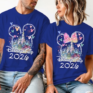 K&ouml;nnte beinhalten: Zwei blaue T-Shirts mit einem Disney Trip 2026 Design. Das Design zeigt ein Schloss, Feuerwerk und Mickey Mouse Ohren. Ein Shirt hat eine rosa Schleife auf den Ohren. Beide Shirts sind kurz&auml;rmlig.