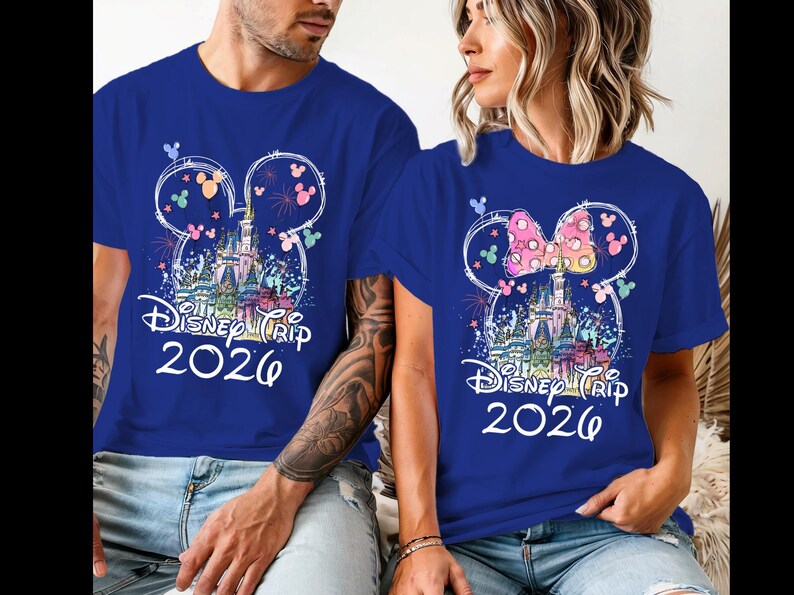 K&ouml;nnte beinhalten: Zwei blaue T-Shirts mit einem Disney Trip 2026 Design. Das Design zeigt ein Schloss, Feuerwerk und Mickey Mouse Ohren. Ein Shirt hat eine rosa Schleife auf den Ohren. Beide Shirts sind kurz&auml;rmlig.