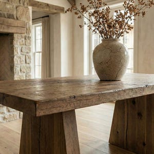 Puede incluir: Una mesa rústica de madera con un acabado envejecido. La mesa tiene una gruesa tapa rectangular y patas anguladas y robustas. Un jarrón decorativo con ramas secas descansa sobre la mesa. El jarrón es de color beige claro con una textura agrietada.