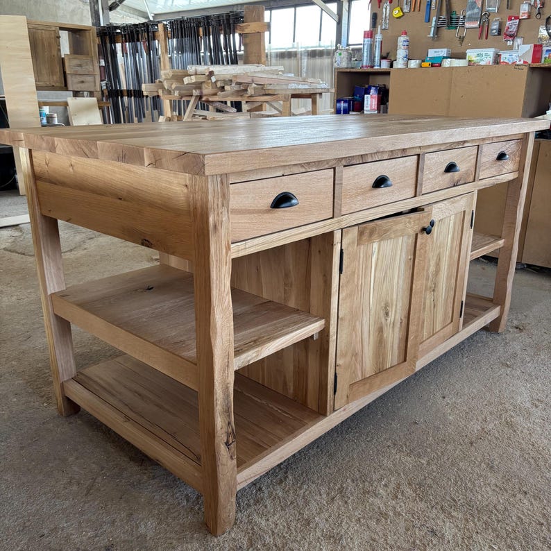 May include: A large rectangular kitchen island crafted from light brown natural wood. The unit features a thick solid wood countertop, three upper drawers with black metal pulls, a central cabinet with two doors, and open shelving on the sides.