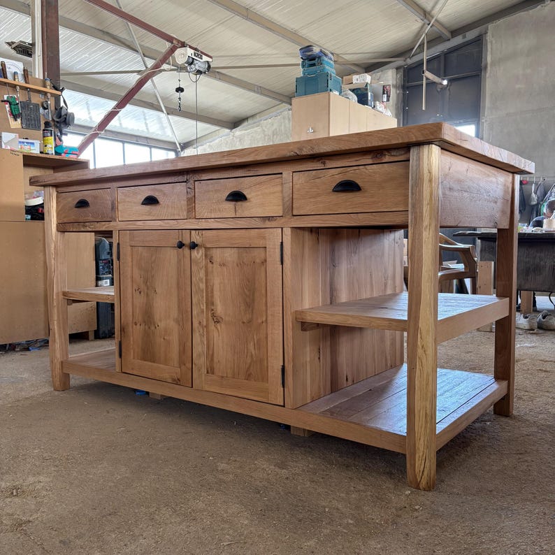 May include: A large rectangular kitchen island crafted from light brown wood with a thick countertop. It features four upper drawers with black semi-circular pulls, two central cabinet doors with small black knobs, and open side shelving units.