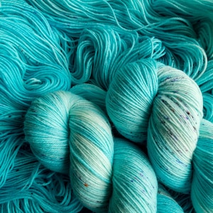 May include: Close-up of a skein of teal blue yarn with white and speckles of colour throughout. The yarn is soft and fluffy, perfect for knitting or crocheting.