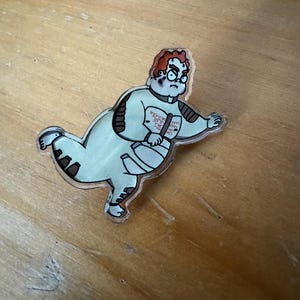 May include: A clear acrylic pin featuring a cartoon character with red hair, wearing a white jumpsuit and glasses. The character has a grumpy expression and is running.