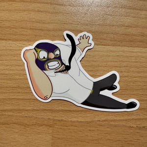 Puede incluir: Una pegatina de dibujos animados de una persona que lleva una máscara de lucha libre morada y dorada, una camisa blanca y pantalones negros. La persona está tumbada de espaldas con los brazos y las piernas extendidos.