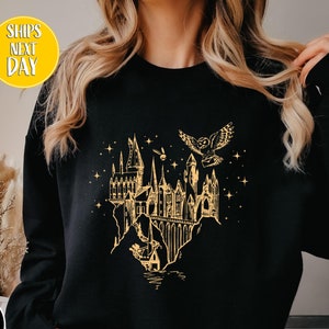 May include: Black sweatshirt featuring a gold illustration of a castle, an owl, and stars. The design is a detailed line drawing. The sweatshirt has a crew neck and long sleeves. The text "BLACK" is in a black circle.