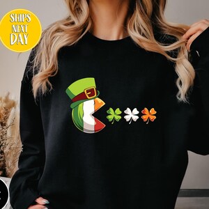 May include: Black sweatshirt with a green leprechaun wearing a hat eating a shamrock cookie. The leprechaun is surrounded by three shamrocks in green, white, and orange.