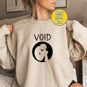 May include: Sand-colored sweatshirt with the word "VOID" above a black circle containing a silhouette of a person in a fetal position. The sweatshirt has long sleeves and a crew neck.
