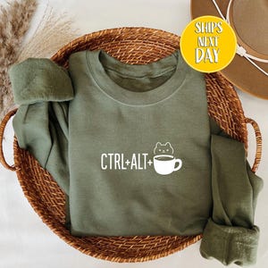 May include: An olive green sweatshirt with the text "CTRL+ALT+" and a cat and coffee cup graphic. The sweatshirt is folded in a woven basket with dried decorative plants and a brown hat.