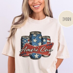 May include: A white t-shirt with a vintage-style graphic of three red, white, and blue American flag-themed cans with the text "Ameri Can" in a red banner.