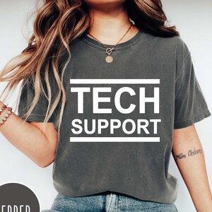 May include: A gray t-shirt with the text "TECH SUPPORT" printed in white on the front. The shirt is worn by a person with long brown hair and a tattoo on their arm.