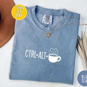 May include: A blue jean colored t-shirt with the text "CTRL+ALT+" followed by a cat and coffee cup graphic. The shirt has a crew neck and short sleeves. A gold necklace is draped across the shirt.