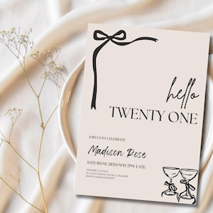 May include: A beige invitation card with black text and a black bow design. The text reads "hello TWENTY ONE" and includes event details. The card is on a white plate with floral accents.