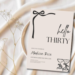 Puede incluir: Una tarjeta de invitaci&oacute;n de color melocot&oacute;n con texto negro y un dise&ntilde;o de lazo negro. El texto dice "hello THIRTY" e incluye detalles del evento. Dos copas de champ&aacute;n est&aacute;n ilustradas en la esquina inferior derecha. La tarjeta est&aacute; sobre un plato blanco y una tela crema.