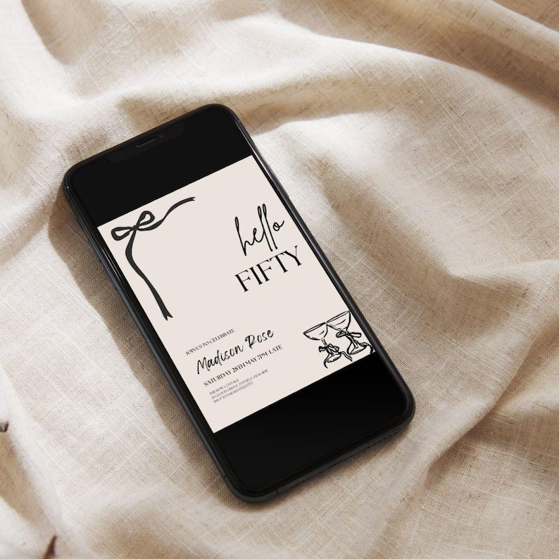 May include: A black smartphone displays a digital invitation with the text "hello FIFTY." The invitation features a bow, two champagne glasses, and the name "Madison Rose." The background is a light beige color.