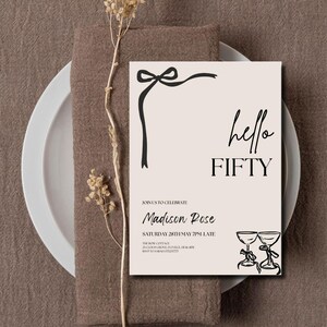 May include: A beige invitation card with the text "hello FIFTY" in black script. The card features a black ribbon design and two champagne glasses. The invitation is set on a white plate and a brown napkin.