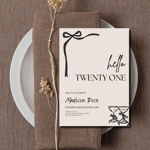 May include: A beige invitation card with black text that reads "hello TWENTY ONE". The card features a black bow and two champagne glasses. The invitation is on a white plate and brown napkin.