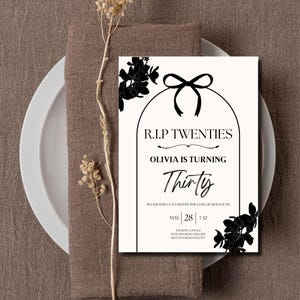 May include: A cream-colored invitation card with black floral accents and text. The text reads "R.I.P Twenties, Olivia is turning Thirty." The card is placed on a brown napkin and white plate, with dried flowers.