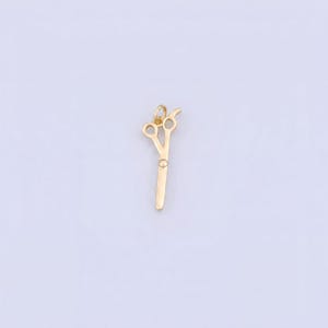 May include: A small, gold-colored metal charm shaped like a pair of scissors. The charm features two circular finger loops at the top and a single, straight blade extending downward, with a small jump ring attached to the top for hanging purposes.