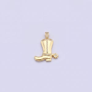 May include: A gold-colored pendant in the shape of a cowboy boot. The boot has a spur and is embellished with small, clear gemstones. The bail and top of the boot are also adorned with gemstones. The pendant is set against a light purple background.