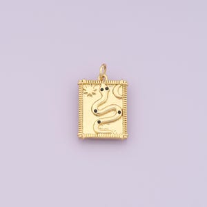 Pode incluir: Pingente dourado em forma retangular. Apresenta um design de cobra estilizado com detalhes em pontos pretos, um sol e uma lua crescente. As bordas têm uma borda texturizada e um pequeno laço na parte superior para uma corrente.