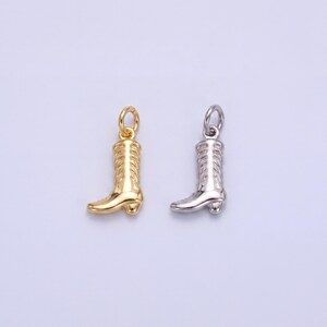 May include: Two small charms shaped like cowboy boots. One is gold-colored, and the other is silver-colored. The boots have detailed stitching and a loop at the top for attaching to a necklace or bracelet. The charms are on a light purple background.