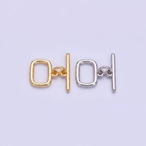 Op de afbeelding: Twee tuimelsluitingen, één goudkleurig en één zilverkleurig, worden getoond tegen een lichte achtergrond. Elke sluiting heeft een rechthoekige lus en een staaf voor sluiting, verbonden door kleine decoratieve elementen.