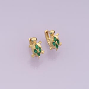May include: A pair of gold-colored hoop earrings, each shaped like a turtle. The turtles have green gemstone accents on their shells and eyes. The earrings are displayed against a light purple background.